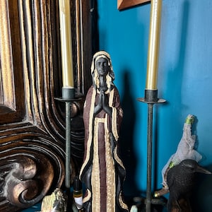 May include: A dark wood framed mirror with ornate carvings, a black figurine of a woman in a long robe with her hands clasped in prayer, two tall white candles in black candle holders, and a black raven figurine on a wooden base.