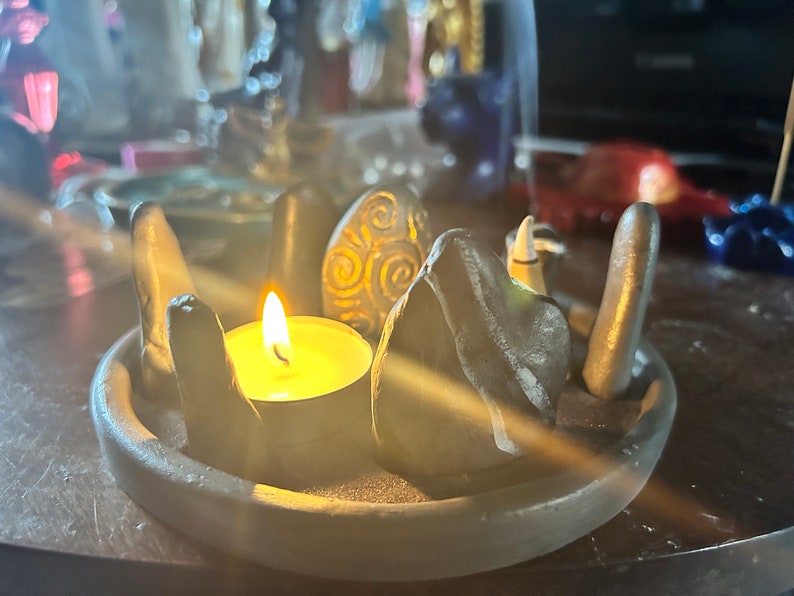 May include: A circular grey clay tray holding a lit tea light candle surrounded by several upright grey stones. One stone features a carved spiral pattern. The arrangement mimics a stone circle, with the candle flame casting a warm glow.
