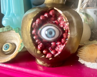 May include: A gold pomegranate with a red interior and a large blue eye peering out. The pomegranate is sitting on a pink surface.