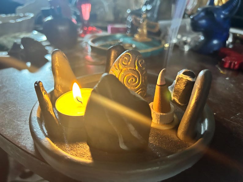 May include: A circular grey ceramic tray holding a lit yellow tea light candle, several smooth dark grey stones, one stone with a carved spiral pattern, and a small conical incense burner emitting a thin trail of smoke into the air.