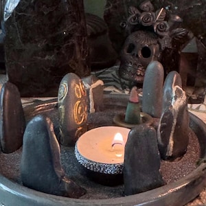 May include: A circular grey tray filled with dark sand containing several upright smooth grey stones arranged in a circle. One stone features gold spiral carvings. A lit white tea light candle with a silver glitter rim and a small incense cone sit inside.