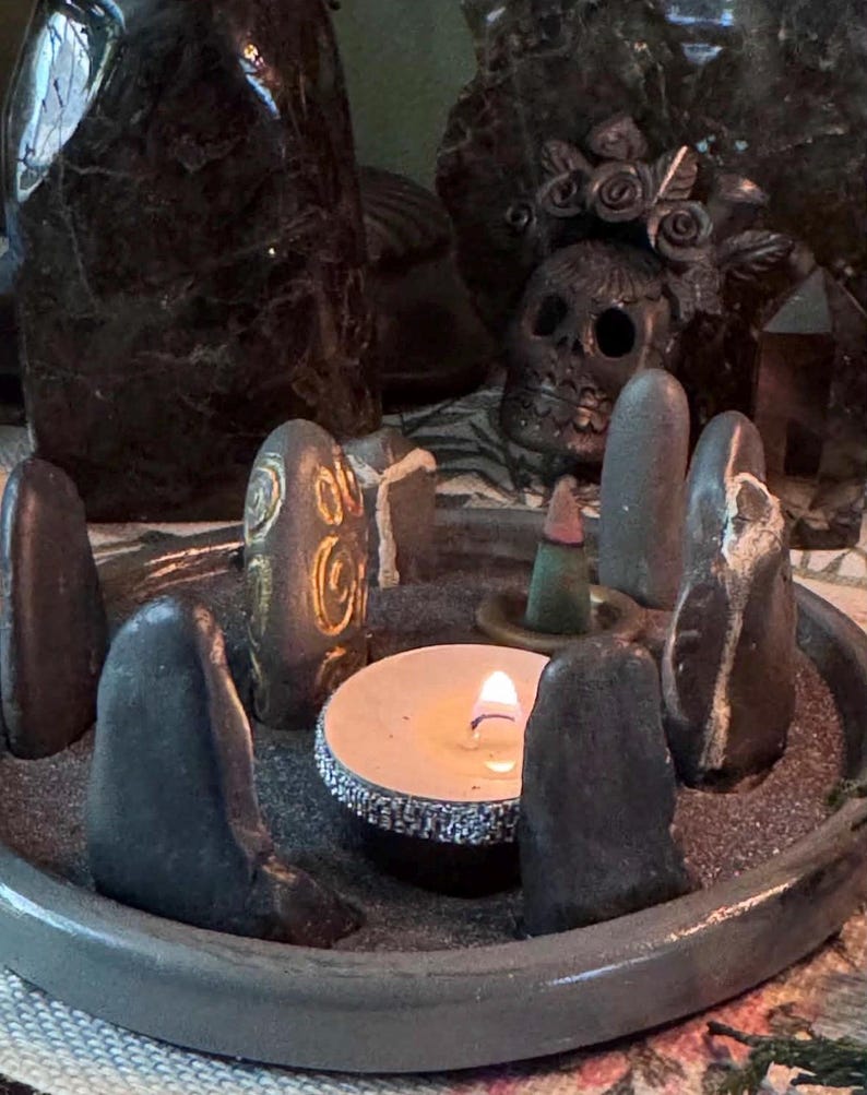 May include: A circular grey tray filled with dark sand containing several upright smooth grey stones arranged in a circle. One stone features gold spiral carvings. A lit white tea light candle with a silver glitter rim and a small incense cone sit inside.