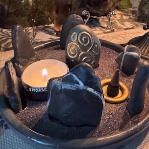 May include: A circular dark grey tray filled with black sand, containing several smooth dark stones, one of which features a gold spiral design. A lit tea light candle in a silver holder and a small incense cone on a gold ring are also present.