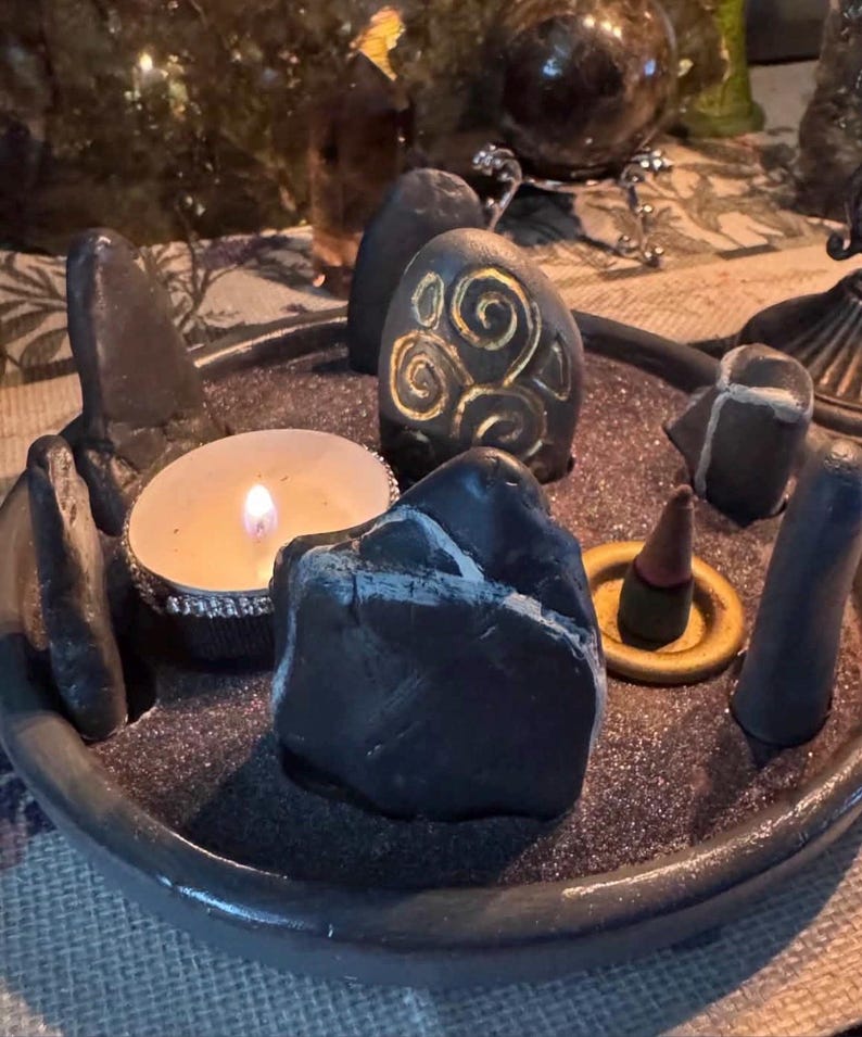 May include: A circular dark grey tray filled with black sand, containing several smooth dark stones, one of which features a gold spiral design. A lit tea light candle in a silver holder and a small incense cone on a gold ring are also present.