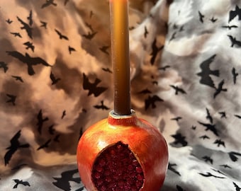 May include: A black candle burning in a red pomegranate-shaped candle holder. The holder is decorated with gold paint and has a detailed design of pomegranate seeds.