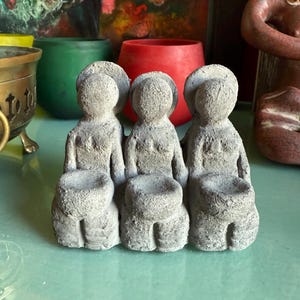 以下が含まれることがあります： 足を組んで座っている人の灰色のコンクリート製の置物3つ。それぞれの置物には、膝の上に小さな丸い窪みがある。