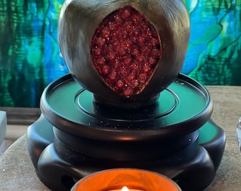 May include: A dark brown ceramic pomegranate with a red glitter interior sits on a black wooden pedestal. A small copper candle holder with a lit candle is in the foreground.