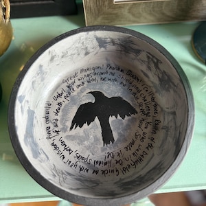 May include: A handcrafted ceramic bowl featuring a black raven design at its centre. The interior is painted in grey and white tones, with handwritten text encircling the raven. The rim of the bowl is dark grey.