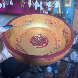 May include: A wooden bowl with a gold interior and a red rim. The bowl features a painted pomegranate in the center and text around the edge. The text includes the words "Persephone" and "sacred hour".