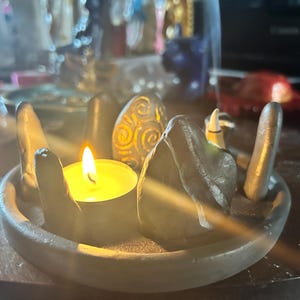May include: A circular grey clay tray holding a lit yellow tea light candle surrounded by several upright grey stones. One stone features a carved spiral pattern. The arrangement mimics a stone circle, with the candle flame casting a warm glow.