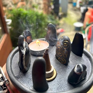 May include: A circular grey tray holding several smooth black stones, a small lit tea light candle, and a cone-shaped incense burner. One stone features gold spiral carvings, while others have white mineral veins. Wisps of smoke rise from the incense.