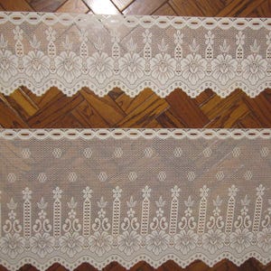 May include: Two strips of off-white lace with floral and geometric patterns. The top strip has a scalloped edge with large flower designs. The bottom strip features a smaller, more delicate pattern with repeating floral and dot motifs. The lace is set against a wooden background.