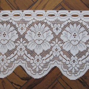 May include: Close-up of white lace trim with a floral design. The lace features a repeating pattern of large flowers and smaller floral accents, with a scalloped edge. The background is a dark wood surface.