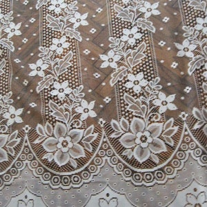 May include: White lace curtain with a floral pattern. The lace has a scalloped edge and a repeating pattern of small flowers and leaves.