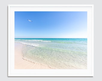 May include: A white sandy beach with clear turquoise water and a single seagull flying in the blue sky.