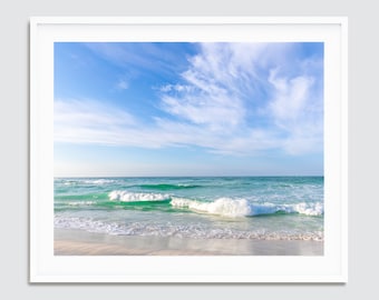 May include: A framed print depicting a beach scene. The image shows a bright blue sky with wispy clouds above turquoise water. White waves break on a sandy shore, creating a tranquil coastal landscape.