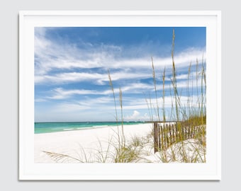 May include: A framed photograph of a beach scene. The image features a white sandy beach, turquoise water, and a blue sky with wispy clouds. Tall grasses are in the foreground, and a wooden fence is visible on the beach. The photograph is in a white frame.