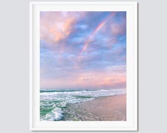 May include: A framed print depicting a beach scene with a rainbow. The sky displays pink, blue, and peach tones, with a double rainbow spanning the clouds. The ocean waves are visible, and the sand is a light tan colour.