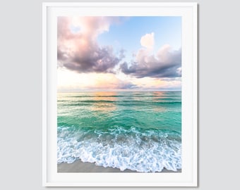 May include: A photograph of a beach scene with turquoise water and white foamy waves crashing on the shore. The sky is a soft blue with fluffy white and pink clouds.