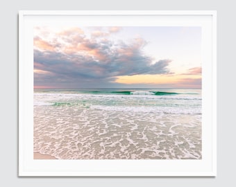 May include: A photograph of a beach scene with a soft, pink and purple sky and white clouds. The ocean is a light blue-green colour with white waves crashing on the shore. The sand is white and the water is clear.