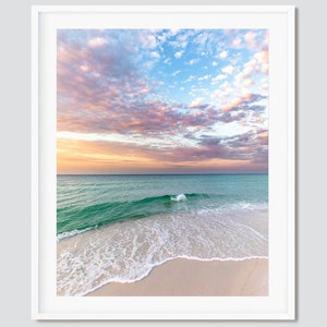 May include: A seascape photograph featuring a sandy beach with foamy waves lapping at the shore. The sky is a vibrant mix of pink, orange, and blue with fluffy clouds.