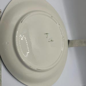 May include: Off-white oval serving platter with a handle. The underside of the platter is visible, showing a glossy finish and some minor imperfections. The handle appears to be made of metal.