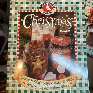 Puede incluir: Una cubierta de libro de cuadros verdes y rojos con el título "Christmas" y las palabras "Gooseberry Patch" en un círculo rojo con una ilustración de un ganso. El libro se titula "Christmas Book 2" y presenta una foto de dos frascos llenos de galletas y una galleta en forma de estrella encima de un frasco. El texto debajo de la foto dice "Yummy holiday recipes, easy-to-make gifts & fun decorating ideas".