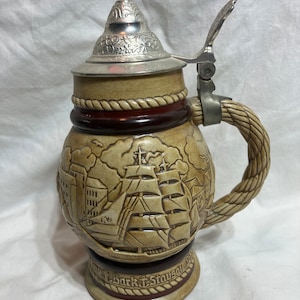 May include: A ceramic beer stein with a brown and white design of a sailing ship and a city skyline. The stein has a metal lid with a hinged handle and a rope-like handle. The base of the stein reads "The Bark Staysail."