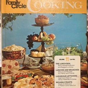 May include: A vintage cookbook titled "Family Circle Illustrated Library of Cooking" with a cover featuring a tiered serving stand laden with food. The cover is orange and blue, with text including "Volume 11" and various food-related topics.