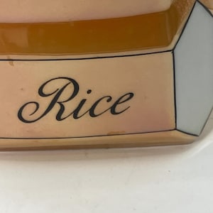May include: A ceramic kitchen canister featuring a cream-colored base with a geometric, faceted design. The word Rice is printed in a black, elegant script font on the front panel, which is framed by thin black lines against a light tan surface.