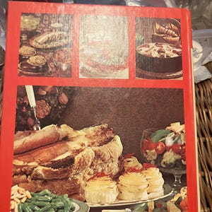 May include: A vintage cookbook with a red cover featuring images of various dishes. The cover displays a collage of food photography, including a roast, vegetables, and desserts. The cookbook's design evokes a retro aesthetic.