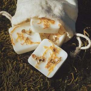 May include: A white canvas bag filled with small, square, white wax melts. The wax melts have dried flowers and herbs embedded in them. The bag is sitting on a bed of green moss.