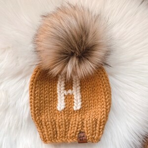 May include: A mustard yellow knitted beanie with a white pom pom and a white letter "H" embroidered on the front. The beanie has a brown leather tag that says "baby knit co."