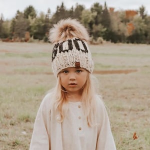 May include: A young person wearing a white and black knitted beanie with a pom pom and the word "GOOD" embroidered on the front. The beanie is worn over a white long-sleeved shirt.