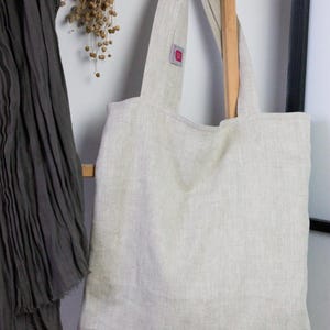May include: A natural linen tote bag with a light beige color and a herringbone weave. The bag has two long straps and a small red label on the front.