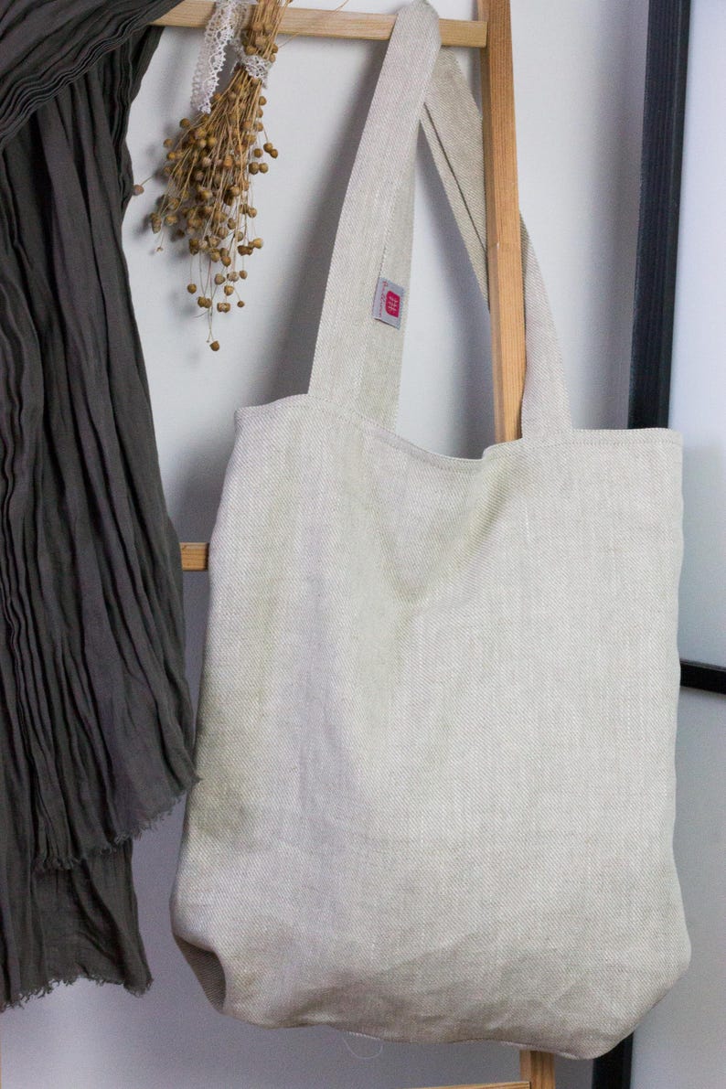 May include: A natural linen tote bag with a light beige color and a herringbone weave. The bag has two long straps and a small red label on the front.