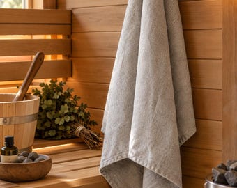 May include: A light beige, textured linen towel hanging from a wooden peg. The fabric displays a natural, woven appearance with visible fibers. The towel is positioned against a light-colored wooden wall, showcasing its simple, functional design.