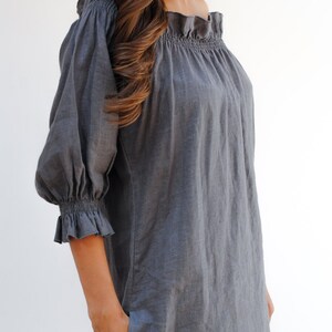 May include: A gray linen dress with a ruffled neckline and 3/4 sleeves. The dress has a relaxed fit and a casual style.