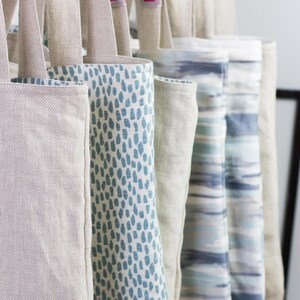 May include: A collection of tote bags with different patterns. The bags are made of a natural linen fabric and have a light blue and white color scheme. The bags are hanging on a rack.