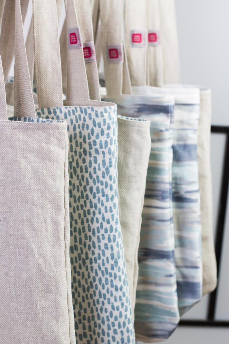May include: A collection of tote bags with different patterns. The bags are made of a natural linen fabric and have a light blue and white color scheme. The bags are hanging on a rack.