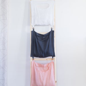 Könnte beinhalten: Ein Satz mit drei Wäschebeuteln, die an einer Holzleiter hängen. Die Beutel sind weiß, grau und rosa.