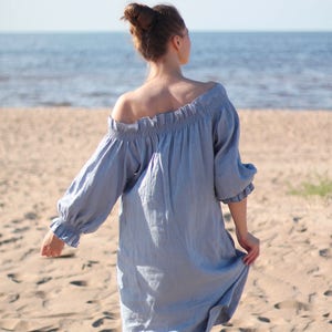 May include: A woman in a light blue off-the-shoulder dress walks on a sandy beach. The dress has a ruffled neckline and long sleeves.