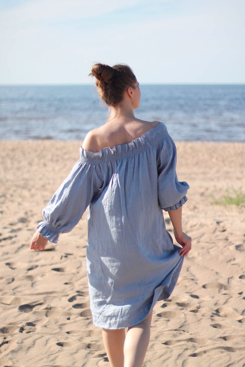 May include: A woman in a light blue off-the-shoulder dress walks on a sandy beach. The dress has a ruffled neckline and long sleeves.