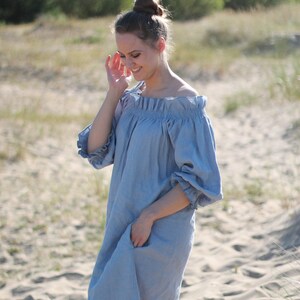 May include: A woman wearing a light blue off-the-shoulder linen dress with puffed sleeves. She is standing on a sandy beach with green trees in the background.