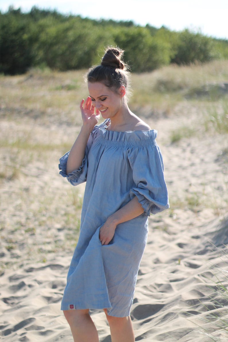 May include: A woman wearing a light blue off-the-shoulder linen dress with puffed sleeves. She is standing on a sandy beach with green trees in the background.