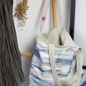 May include: A large, striped tote bag with a light blue, gray, and white watercolor design. The bag has two long, natural linen straps and a small, pink label with the word "Made" on it.