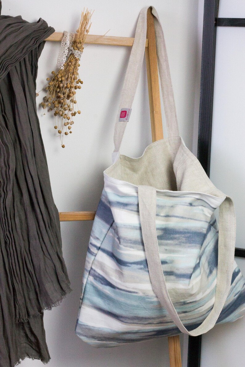 May include: A large, striped tote bag with a light blue, gray, and white watercolor design. The bag has two long, natural linen straps and a small, pink label with the word "Made" on it.