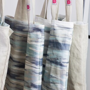 May include: Four tote bags hanging on a rack. Two of the bags are made of natural linen and have a pink logo that says "The Linen Works". The other two bags are made of a blue and white patterned fabric.