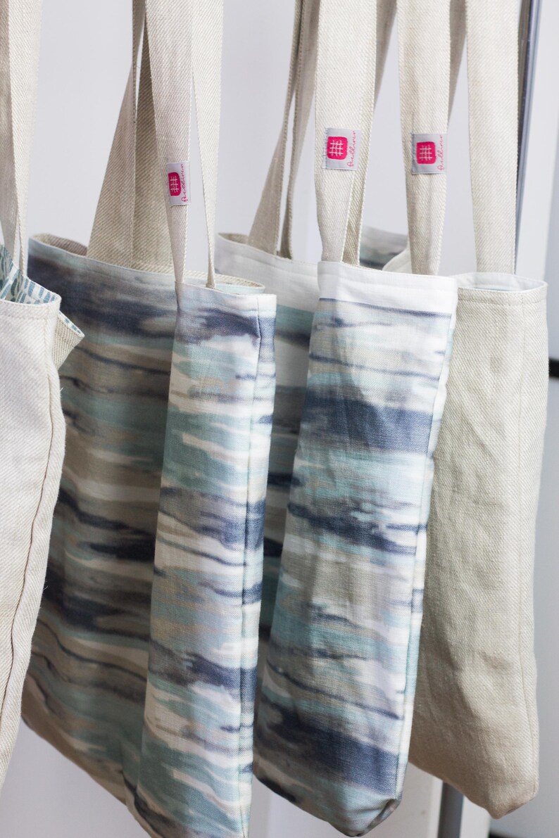 May include: Four tote bags hanging on a rack. Two of the bags are made of natural linen and have a pink logo that says "The Linen Works". The other two bags are made of a blue and white patterned fabric.
