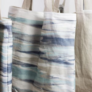 May include: Three tote bags hanging on a white wall. Two of the bags are made of natural linen and have a blue and white striped pattern. The third bag is made of natural linen and has a solid beige color. All three bags have a pink label with the word "Hollown" printed on them.